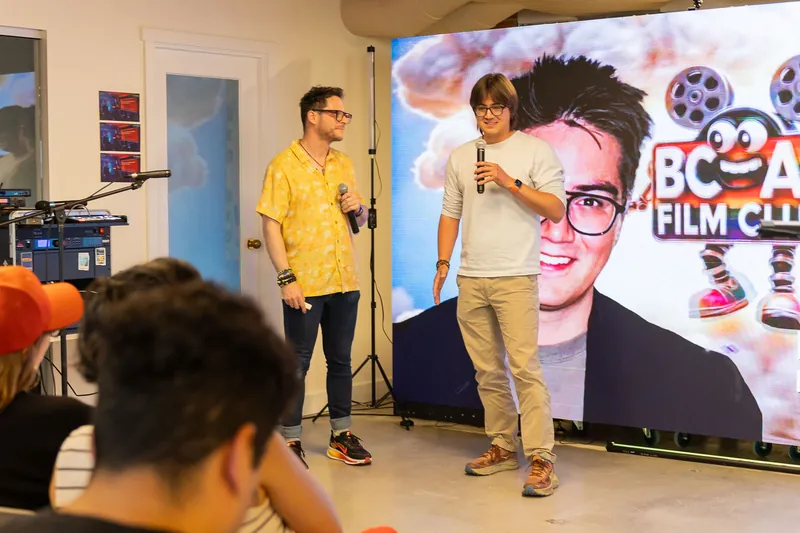 Host handoff and participant presentation beside a projected portrait, photo 178.