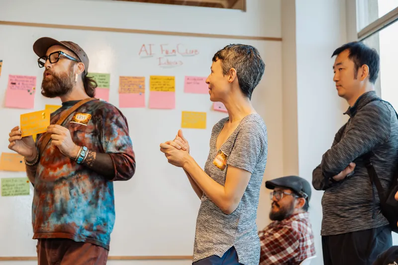 Three participants compare ideas in front of the workshop wall, photo 129.