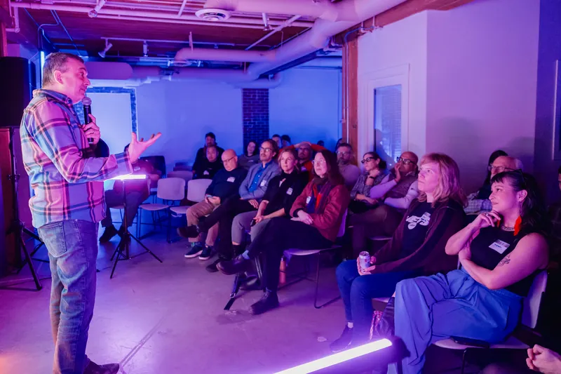 A presenter speaking beside a bright projection with the audience in view.