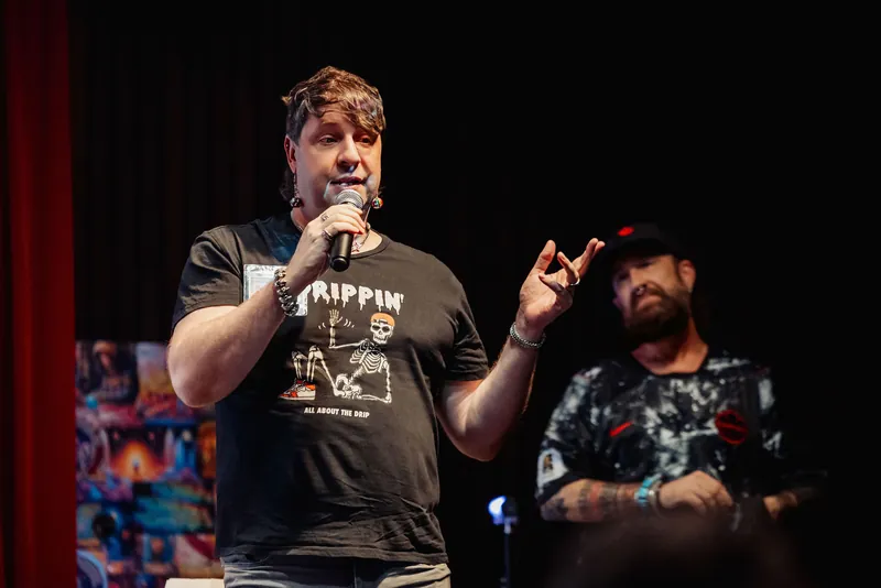 Two presenters share the stage — one addresses the crowd with mic in hand while the other listens in a candid between-beats moment.
