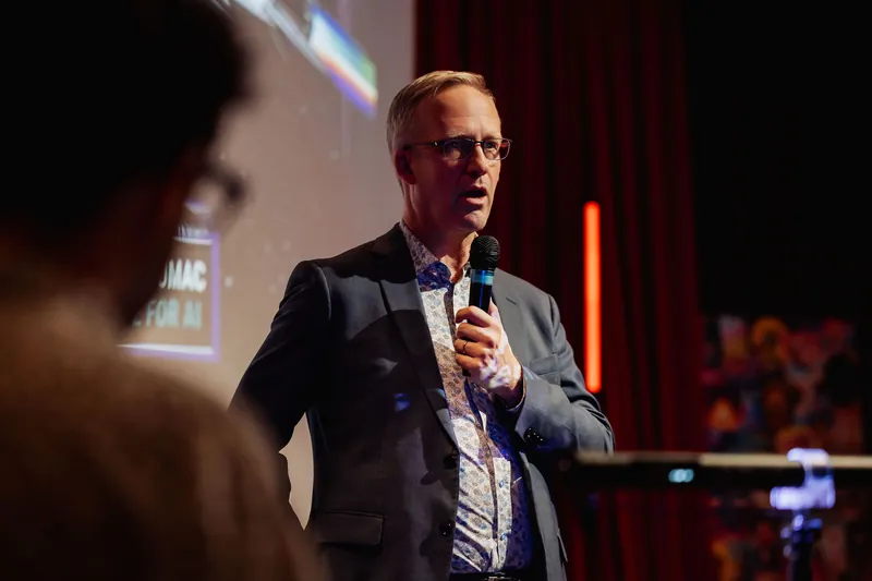 The presenter is framed between a blurred foreground figure and the warm stage lighting, holding the mic and addressing the room. Red curtains and a projected slide are visible in the background, with a vertical lighting rig glowing red at the far right.