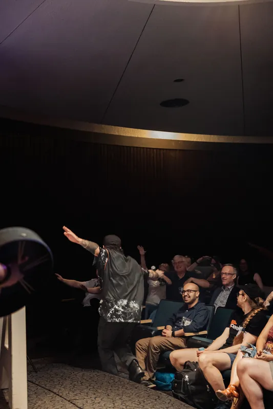 Hands go up across the planetarium as a performer brings the audience to its feet in a high-energy interactive moment.