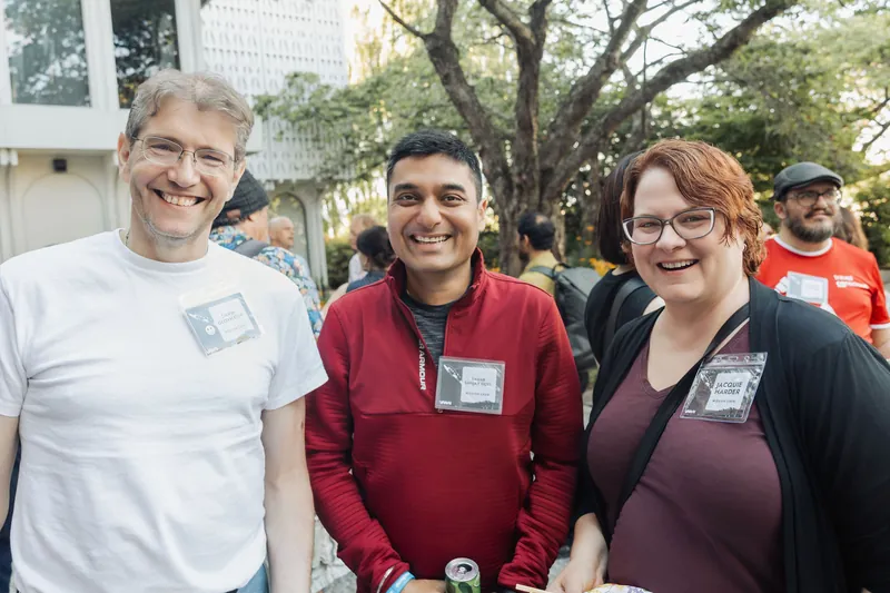 Three attendees smile for the camera at the outdoor plaza reception, drinks in hand.