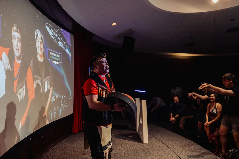 A performer in Indigenous regalia takes the mic beneath the Space Centre's curved dome projection as the audience watches.