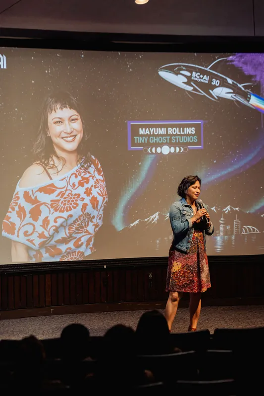 Mayumi Rollins grins mid-talk, the cosmic aurora projection and her speaker card glowing warmly behind her.