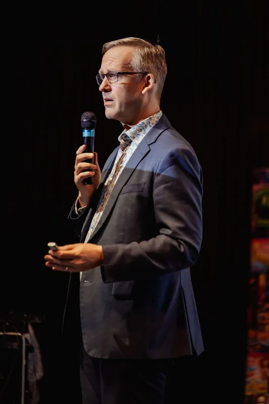 Full-length profile of the speaker against a dark backdrop, mouth open mid-speech with the mic held high. A horizontal stage light bar on a stand glows in the background, the only other light source in the frame.