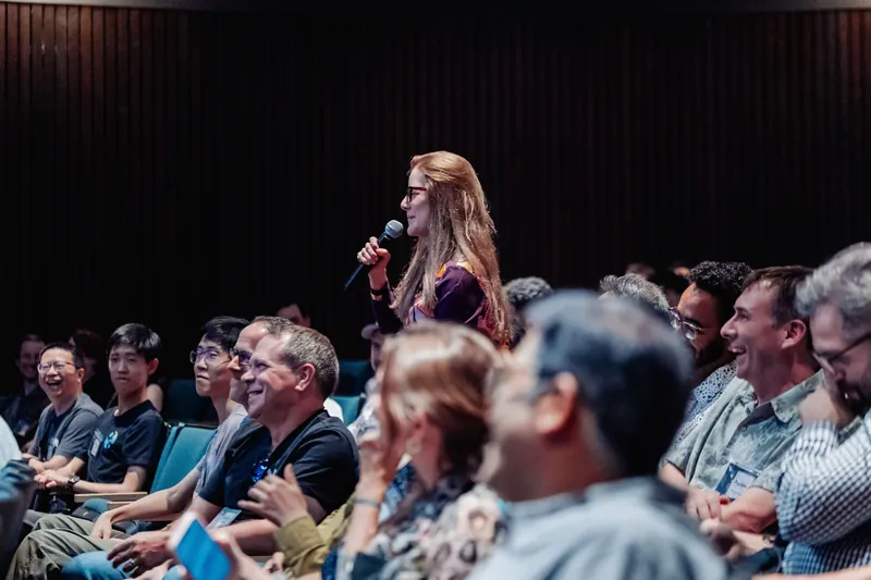 An audience member stands to ask her question as rows of smiling attendees look on in the darkened auditorium.
