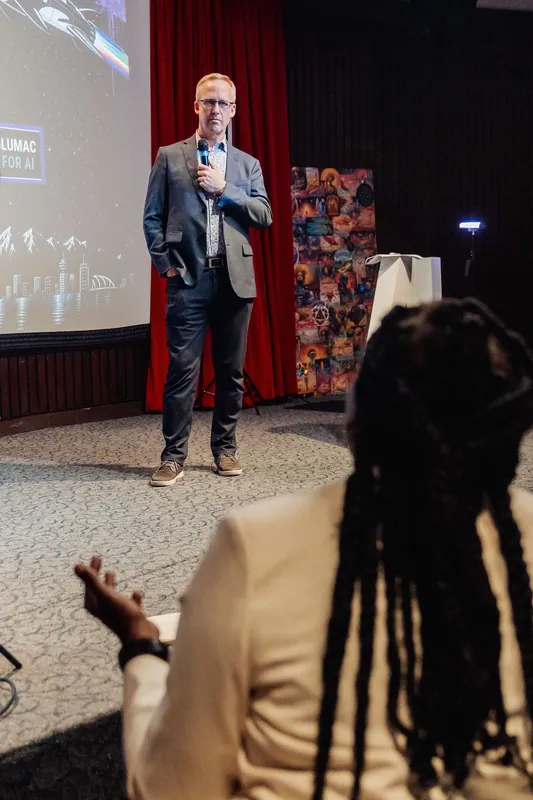 A full-length view of a speaker standing centre-stage, mic raised, with a large projection screen lit behind them. In the foreground an audience member with locs in a cream jacket leans forward, hand lifted slightly. A white podium and a colourful printed banner are visible to the right of the stage.