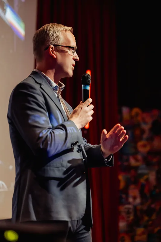 A wider stage view shows the presenter holding the mic and gesturing with one hand slightly raised. A large projected slide is visible in the upper left, and a vibrant decorative backdrop element fills the right side of the frame.