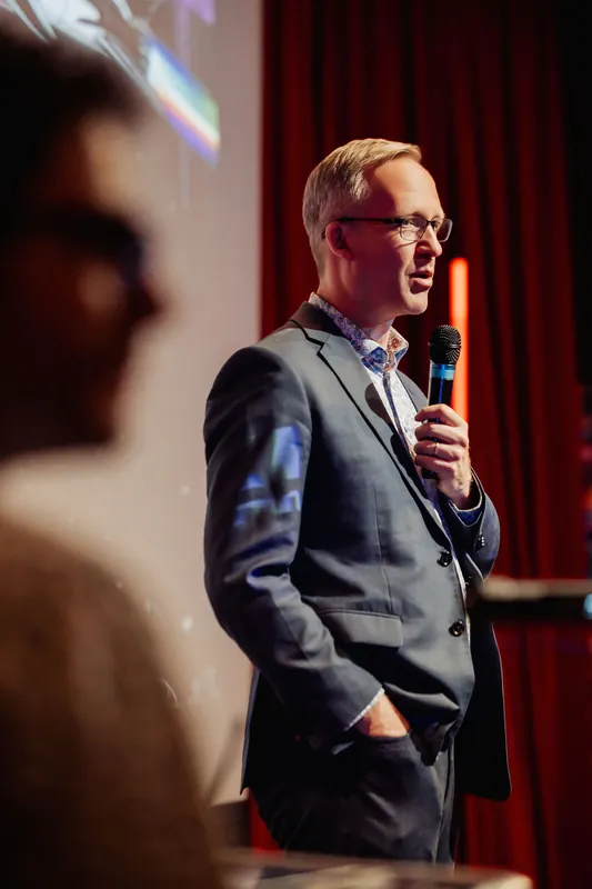 A speaker in a gray blazer and patterned collar holds a handheld mic at chin level, one hand tucked in a pocket, addressing the room in a three-quarter profile. A projected slide glows in the upper background while a blurred audience member fills the foreground, the venue's red walls and warm stage lighting framing the shot.