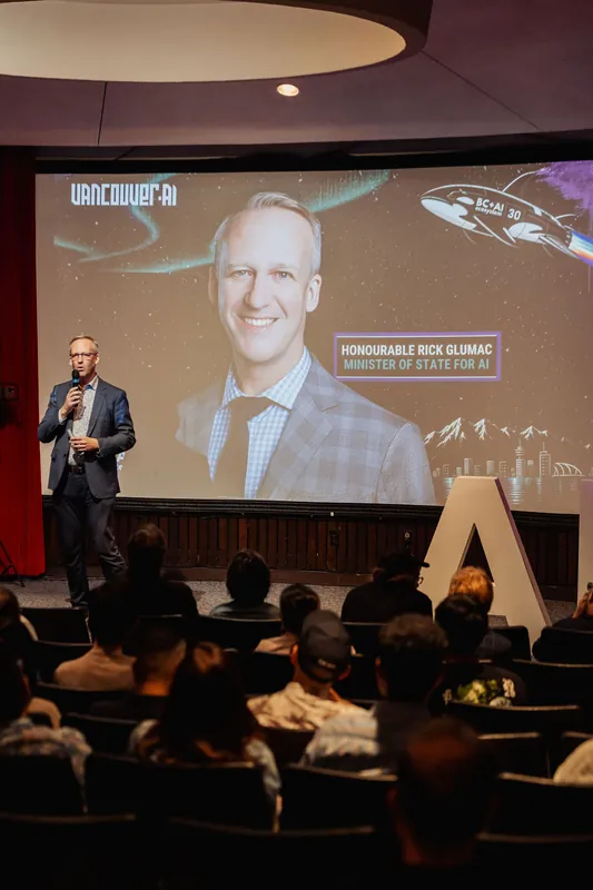 Wide view of the stage with the host at the left holding a mic. A large projection screen fills the back wall with a headshot and event graphics. The audience sits in darkened tiered seating, and tall decorative letters are visible stage right. The circular ceiling of the planetarium is just visible at the top.