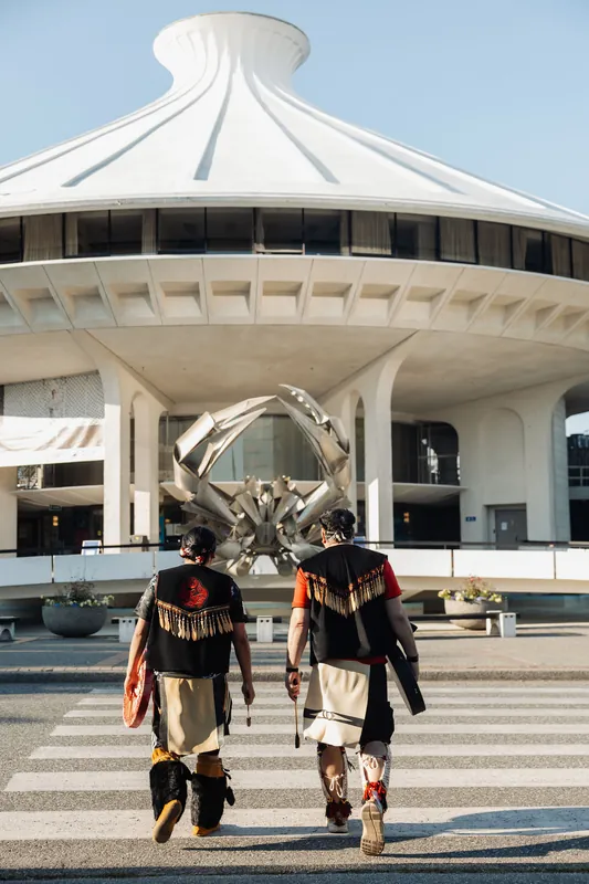 The performers walk side by side across a crosswalk toward the Space Centre's iconic dome and crab sculpture.