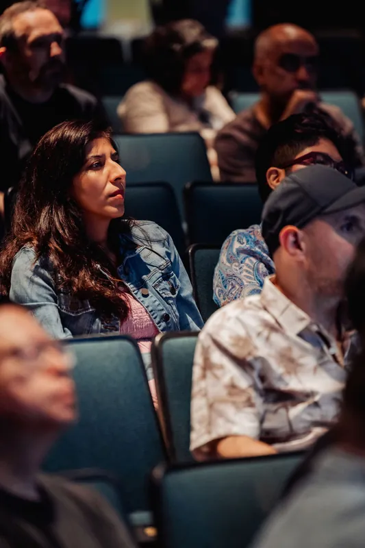 A woman in a denim jacket leans forward to look intently at the stage from her seat in the theater.