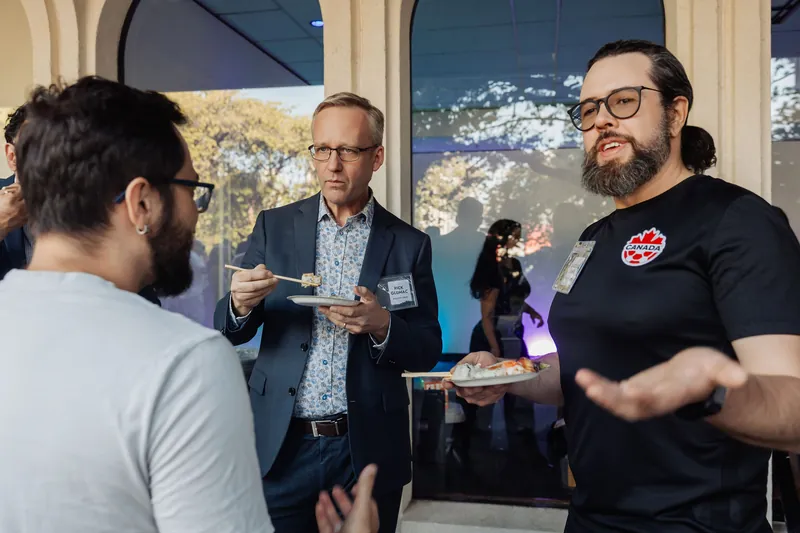 Three attendees in animated conversation over food near an arched window at the evening reception.