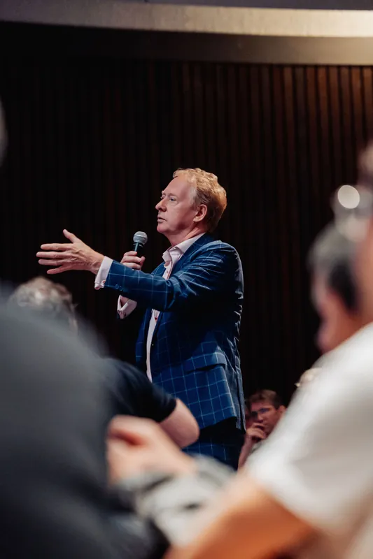 The host in a blue check blazer sweeps one arm toward the crowd while speaking into the handheld mic.