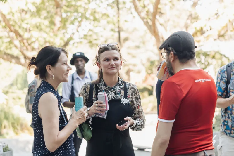 Three attendees laugh together over drinks under dappled trees on the outdoor plaza at golden hour.