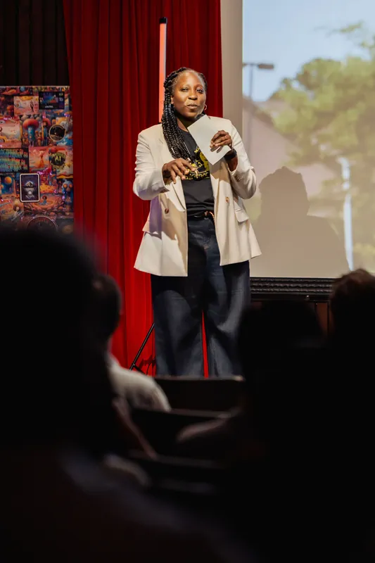 Anthonia Ogundele addresses the seated crowd from the stage, a colorful AI art poster visible to her left.