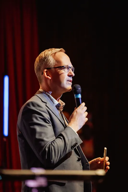 The emcee speaks with quiet intensity, lit by stage light against a red curtain.