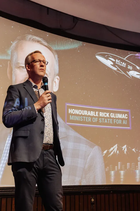 A speaker stands upright on stage, mic at shoulder height, posture composed and slightly turned. A large projected event graphic with a lower-third text overlay fills the curved screen behind them.