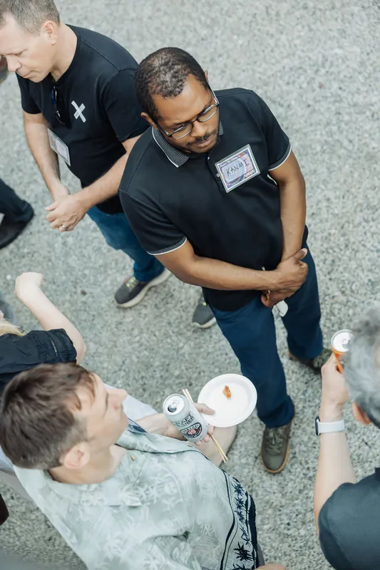A man in a black polo listens intently as the outdoor reception conversation flows around him.