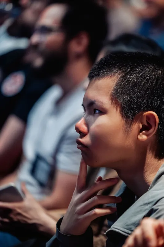 A young attendee listens intently from the theater seats, hand resting at chin.