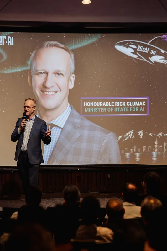 A host stands at the left edge of the stage holding a handheld mic, introducing the evening in front of a large projection screen. The screen displays a prominent headshot portrait alongside event branding. Rows of seated attendees fill the foreground.