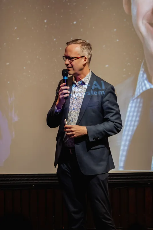 A three-quarter profile of the host mid-speech, mic held at chin height, gaze directed off to the side. The projected slide behind them shows a partial portrait image and branding text softly out of focus.