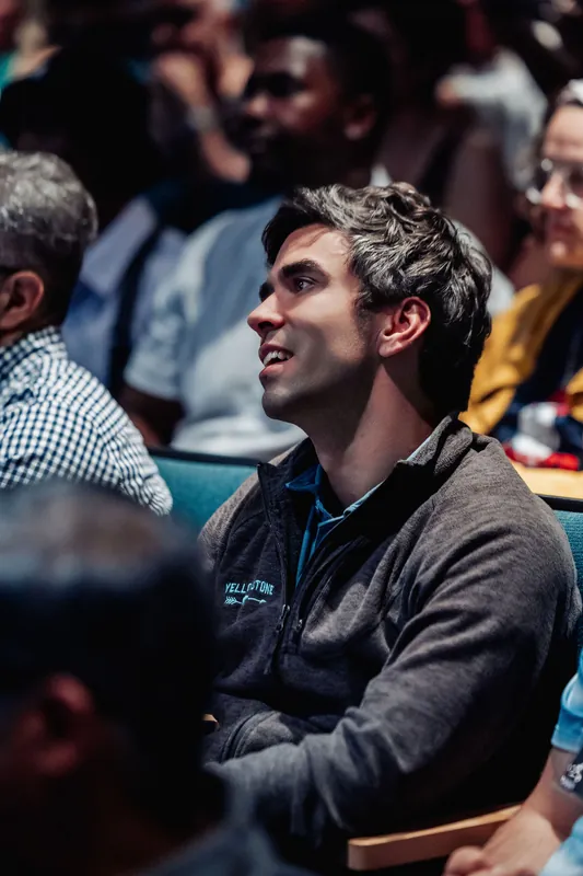 A smiling attendee looks up toward the stage, arms comfortably crossed.
