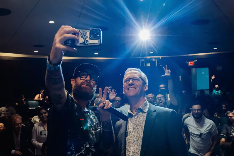 The selfie moment builds as attendees behind raise peace signs and arms, the celebratory energy in the room surging.