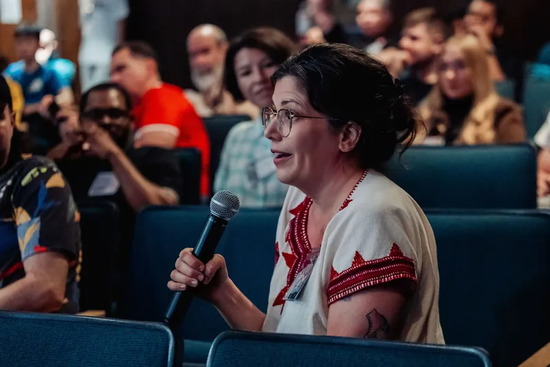 An attendee in a white embroidered top rises with a microphone during Q&A as the crowd smiles warmly behind her.