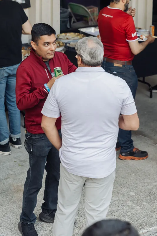 Two attendees exchange conversation at the outdoor reception near the food table, other guests milling about behind them.