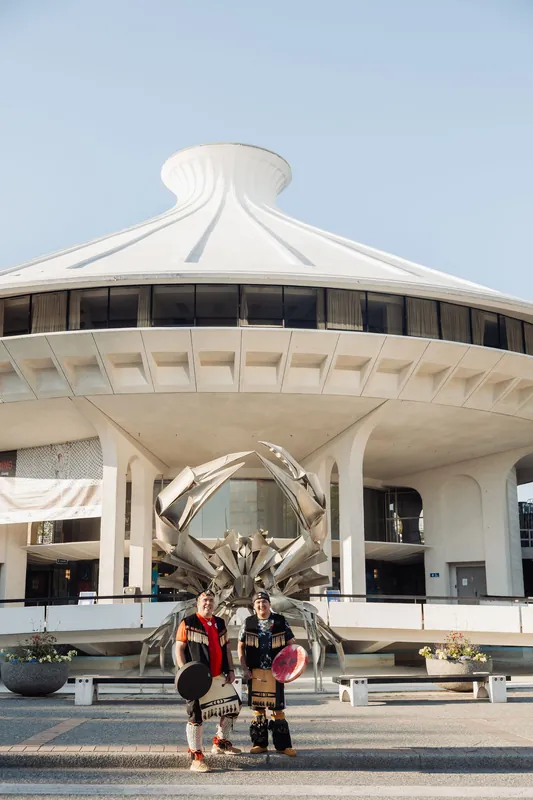 The two performers turn to face the camera, drums in hand, posed before the towering crab sculpture at the Space Centre.