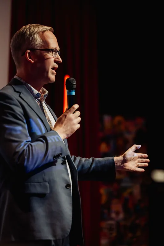 The presenter stands in profile, mic lifted toward their mouth, while the free arm extends out with an open palm. An orange accent light glows behind them against the red curtain, and a colorful backdrop element is visible at the lower right.