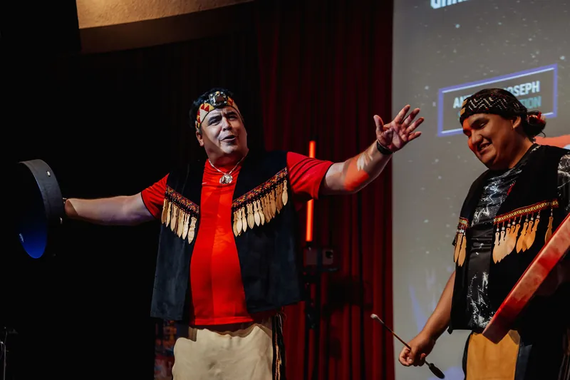 A Squamish Nation performer raises an open hand mid-song as his partner holds a hand drum behind him under stage lighting.