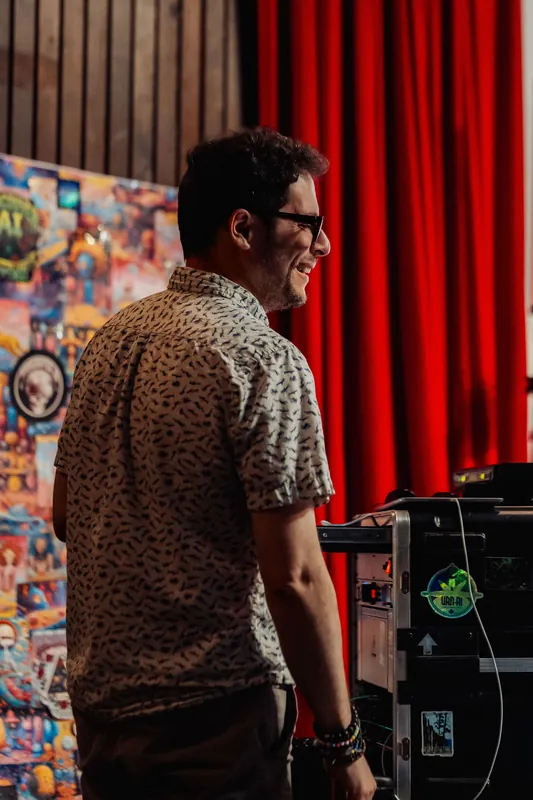 An event organizer at the production desk smiles toward the stage, colorful illustrated art panels visible in the warm backstage light.