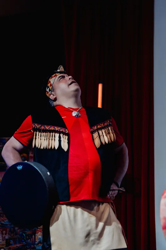 Bathed in warm red stage light, a cultural performer looks skyward with a hand drum during the opening ceremony inside the Space Centre.