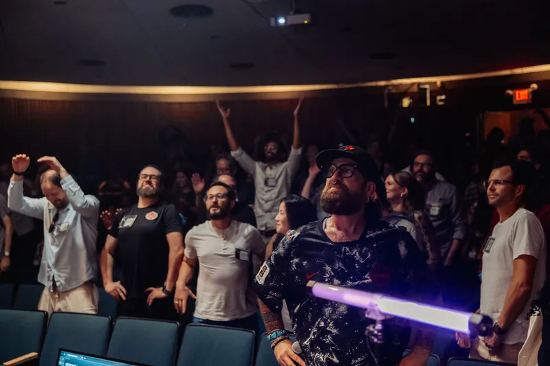 The crowd surges to its feet with arms raised as an attendee waves a glowing lightsaber prop through the air.