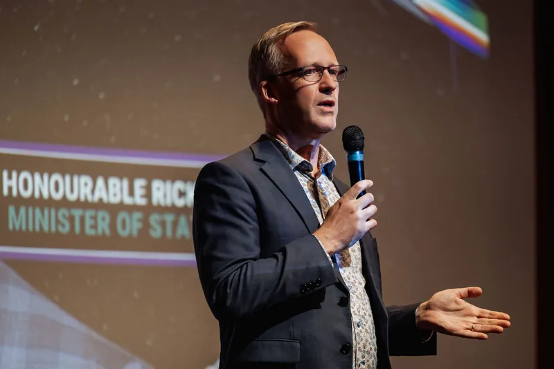 The presenter stands front-facing, microphone held upright, one hand resting near the chest. The projected slide behind them shows a name lower-thirds card, and the room's warm ambient light wraps the stage.