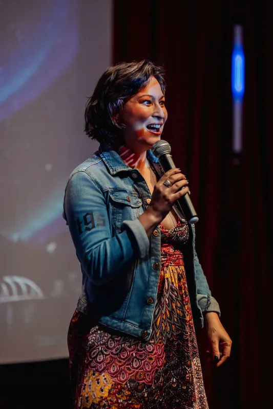 Close-up of Mayumi Rollins, denim jacket catching the stage light, mid-sentence during her Tiny Ghost Studios talk.