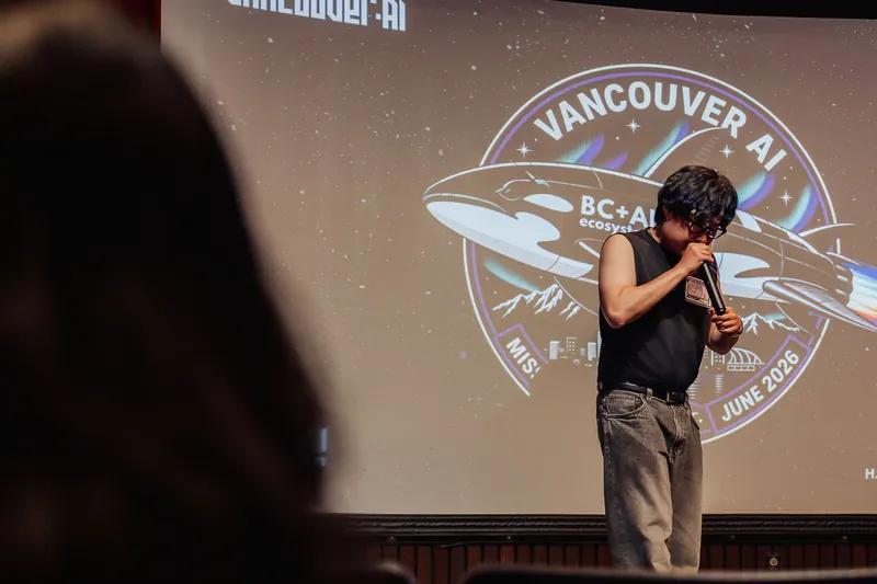 Seen from behind the audience, the emcee stands on stage against the large BC+AI Mission 30 projection.