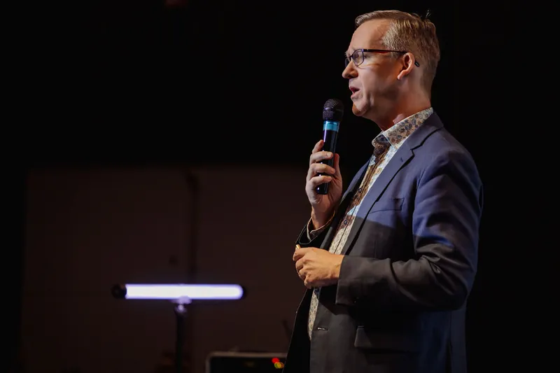 Near-profile view of the speaker in the dark stage space, mic raised, holding a clicker at their side. Colorful artwork or event display panels are visible in the background to the right.