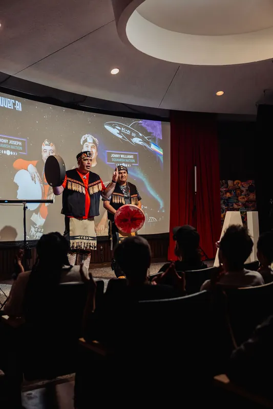 The two Squamish Nation drummers face the seated audience while the Vancouver AI Meetup branding shines on the planetarium screen.