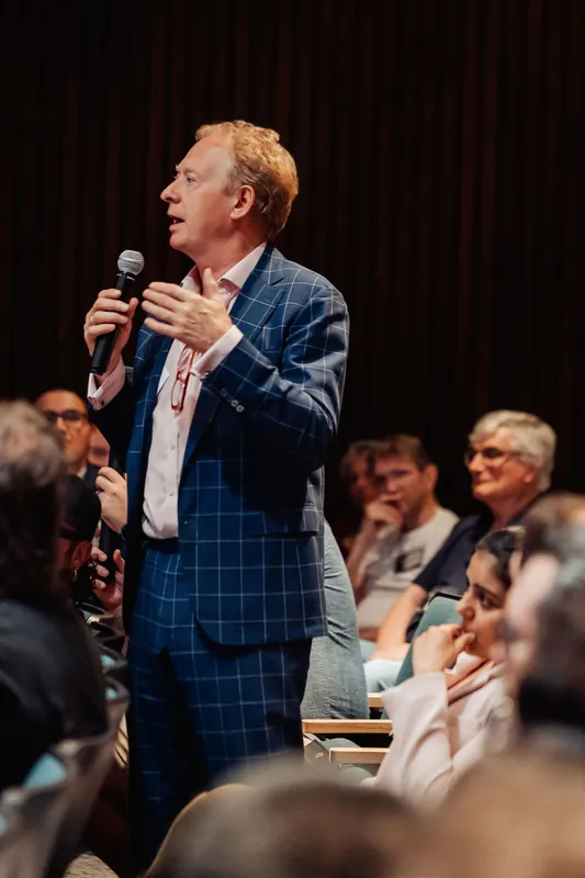 A speaker engages the full auditorium, with rows of attentive attendees stretching behind him.