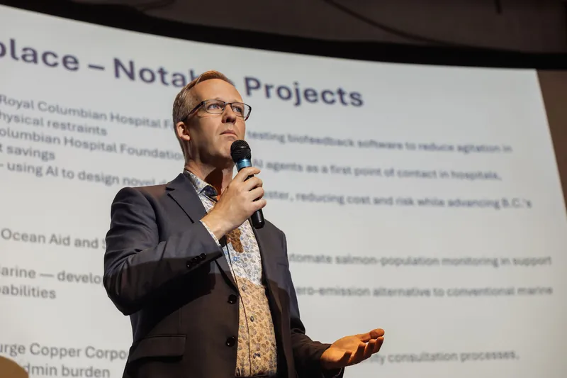 The same presenter stands in three-quarter profile, mic at chin level, other hand extended palm-open toward the audience. A projected slide fills the curved screen behind them.