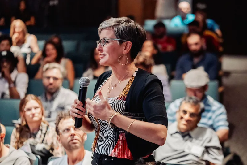 She gestures thoughtfully while speaking, the rows of teal seats and an attentive audience filling the background.