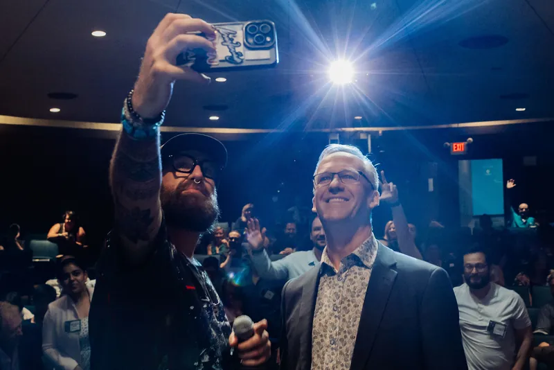 An emcee snaps a selfie with a guest speaker as stage lights blaze and the delighted crowd erupts behind them.