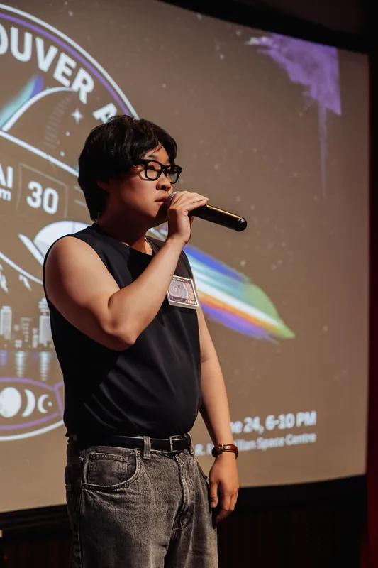 The emcee mid-sentence at the mic, Space Centre event details visible on the projection slide behind them.