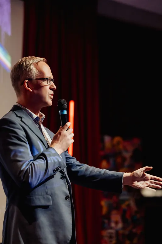 A speaker in a grey blazer and glasses, shown in partial profile, holds a handheld microphone while extending the opposite arm outward with an open palm toward the audience. A colorful projected graphic is visible at the right edge of the frame, and red stage curtains frame the dark background.