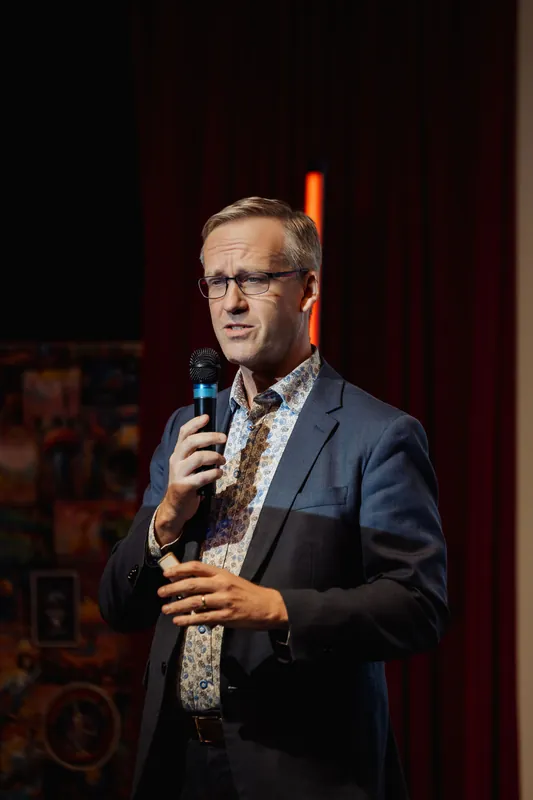 The speaker stands in a dark stage environment, lit from the front by a warm spotlight, mic in hand and looking outward. A horizontal stage light bar sits on a stand behind them in the otherwise unlit space.