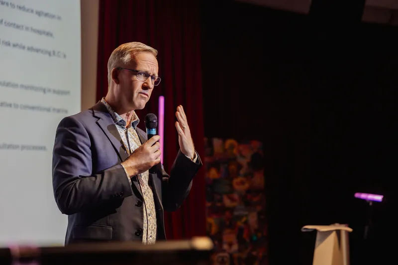Facing more directly toward the camera, the presenter holds the microphone at chest height while raising the other hand palm-out in an emphatic gesture. A projected slide and colorful stage backdrop are visible on either side.